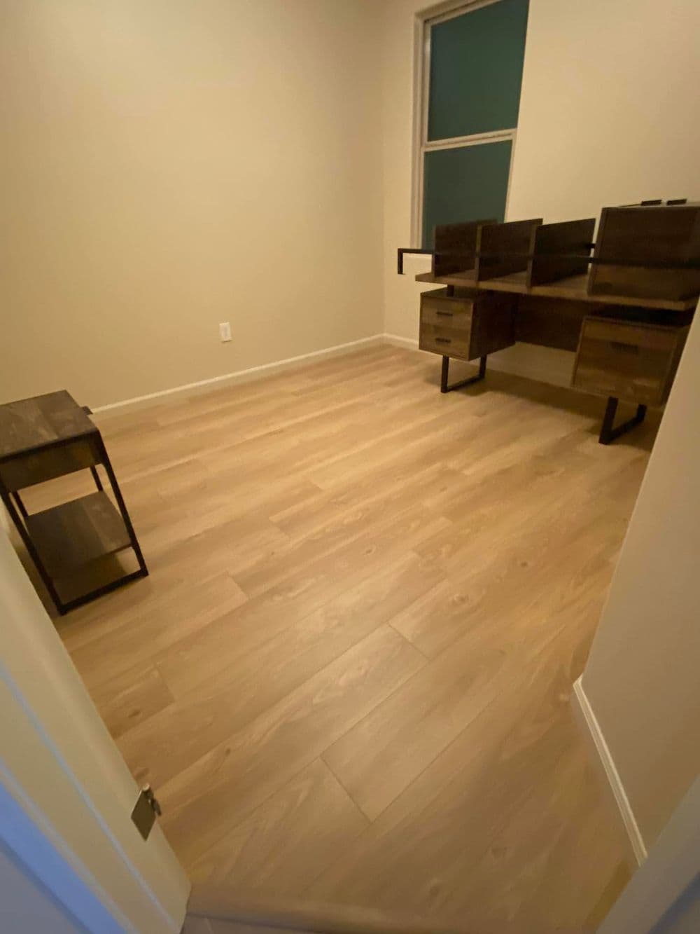 Spacious empty room with light wood floors and modern desk furniture against a neutral wall.
