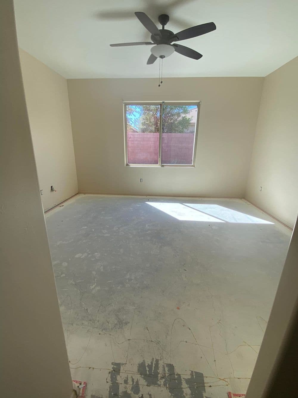 Empty room with beige walls, ceiling fan, and large window letting in natural light.